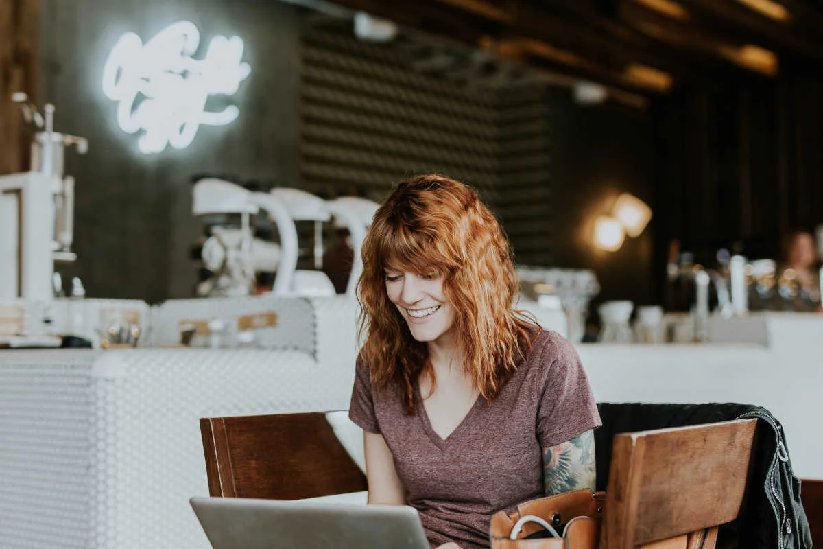 Donna Con Capelli Rossi Sorride Mentre Lavora Al Laptop In Un Caffè Illuminato Da Luci Al Neon.