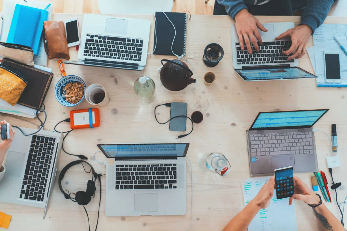 Persone Lavorano A Un Tavolo Con Laptop, Telefoni, Cuffie E Quaderni, Creando Un'atmosfera Produttiva.